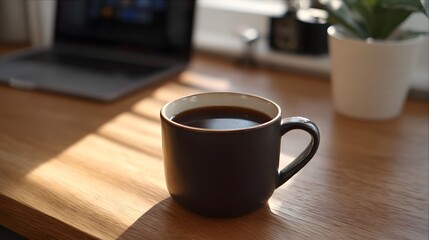 Obraz premium A dark mug of coffee rests on a wooden desk beside a laptop and potted plant dramatically lit by natural morning light