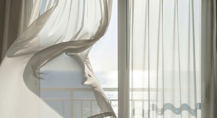 Obraz premium White sheer curtains blowing in the breeze at an open balcony window overlooking the bright sea during a sunny summer day.