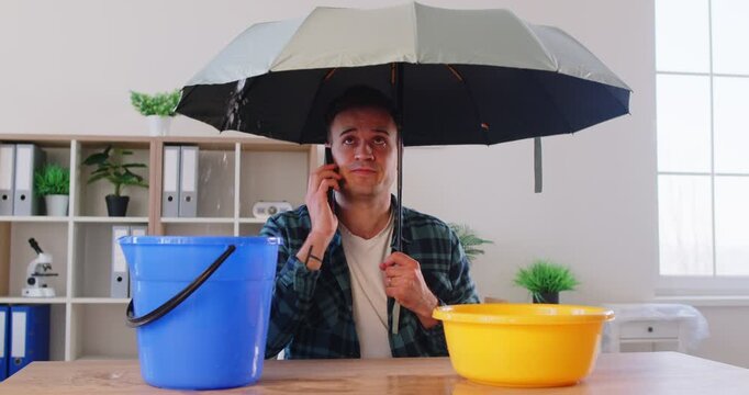 Emergency call to plumbing repair service by man with umbrella due to ceiling leak and broken roof. Worried sad person calling to plumber during home apartment flood, water leaking into buckets