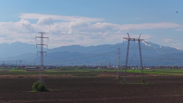 Striking network of high voltage power lines over freshly plowed agricultural fields with hazy snow capped mountains, low altitude slow backward drone shot, 4K