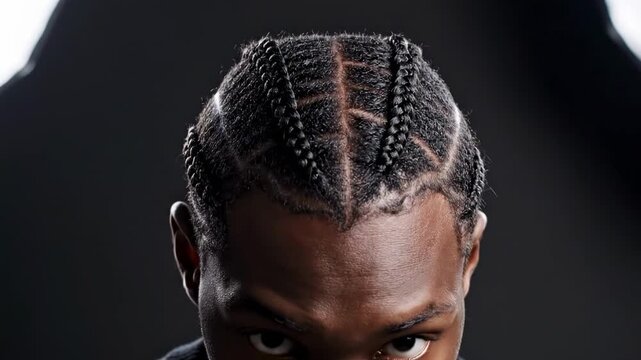 Close studio portrait of a man with cornrows hairstyle, precise scalp pattern, dark backdrop, soft rim light, calm side gaze, clean facial grooming, contemporary masculine beauty editorial style