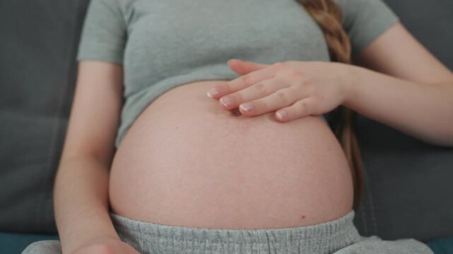 Pregnant white woman massaging baby bump with gentle strokes, manicured nails glide over skin, braided hair visible, relaxed couch pose, serene light, mindful bonding and prenatal selfcare.
