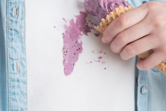 A person is holding an ice cream cone with a visible stain on their shirt. The background shows a simple indoor setting. It is daytime and the person appears to be in a relaxed mood.