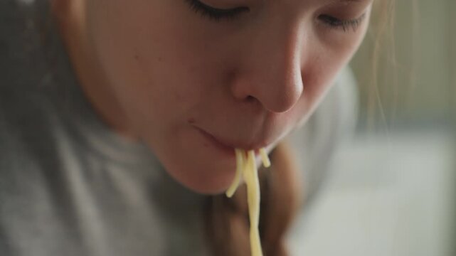 Woman eats noodles. Student enjoys quick meal. Woman in kitchen with braided hair eating noodles happily. Focused student with braided hair relishing comforting meal of long pasta strands