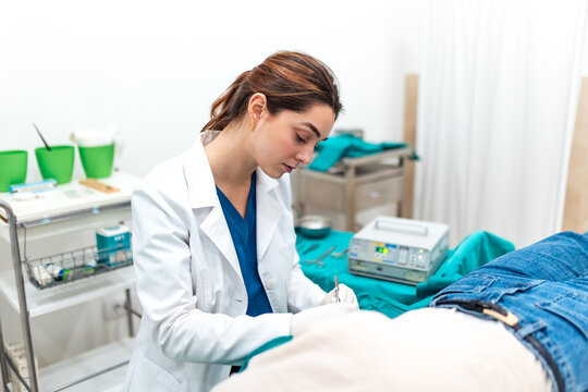 Focused young female surgeon in white coat performing a precise minor surgical procedure on a patient in a medical clinic. Professional healthcare setting with advanced equipment and sterile tools.
