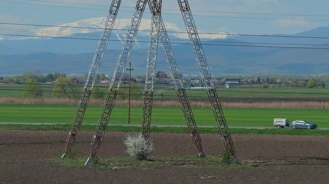 Imposing high voltage transmission tower over agricultural fields and pastures with stunning snow capped mountains, telephoto lens compression slow ascending drone shot, 4K