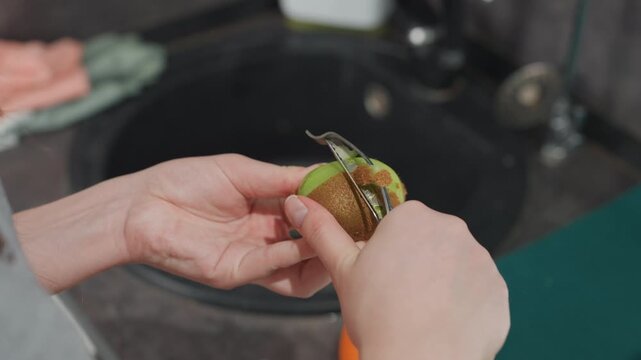 Woman with pregnancy peeling kiwi in kitchen. Expectant woman slices kiwi for healthy snack at home. Pregnant woman is preparing fresh kiwi snack using peeler at home in morning light