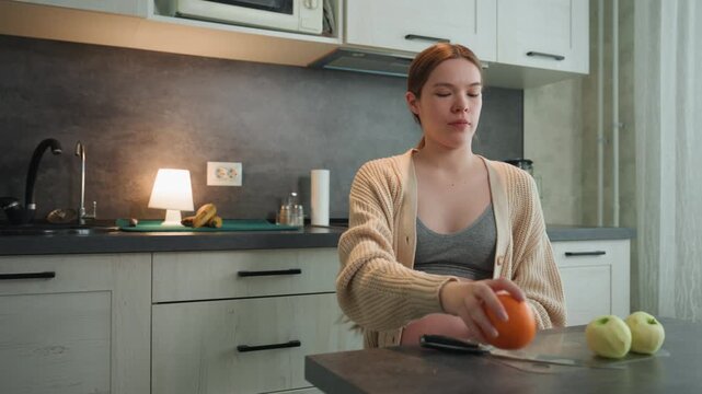 Pregnant white woman in kitchen holding orange and apples, wearing cozy cardigan and bralette, exposed baby bump, soft lamp glow and morning light, smartphone on counter, thoughtful expression