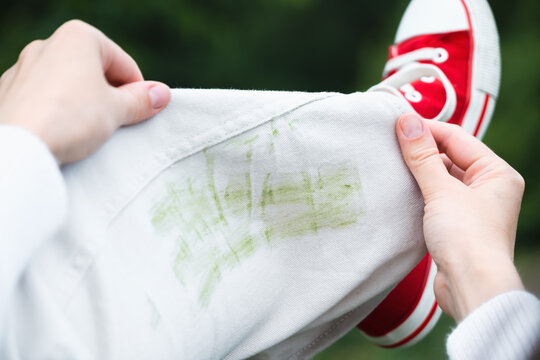 Unrecognizable person demonstrated up their white jeans to show a green stain on the fabric. The individual is wearing red shoes and is outside near trees. Concept of daily stains on clothes. 
