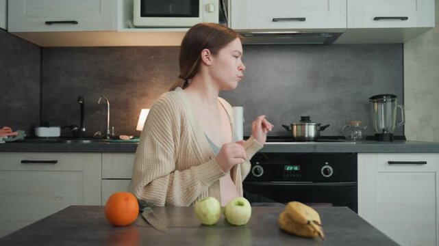Pregnant caucasian woman at kitchen island between fruit and stove tapping orange, glancing at phone, moving to oven and stretching, wearing cozy sweater and bralette, expressive gestures, expectant