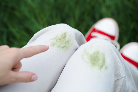 Unrecognizable person demonstrated up their white jeans to show a green stain on the fabric. The individual is wearing red shoes and is outside near trees. Concept of daily stains on clothes. 