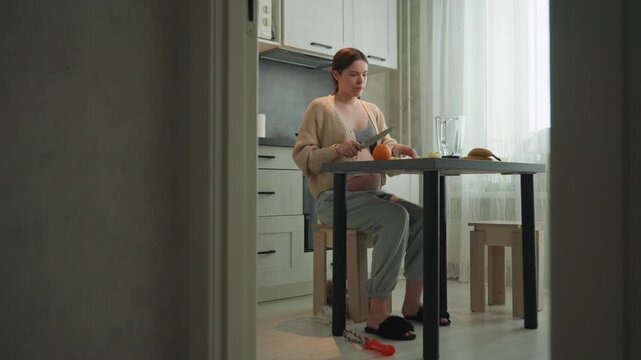 Pregnant white woman sitting at table cutting orange in soft morning light by window. Cozy kitchen with glass jar, stool, slippers and patterned sweater, loose pants, calm introspective mood, gentle