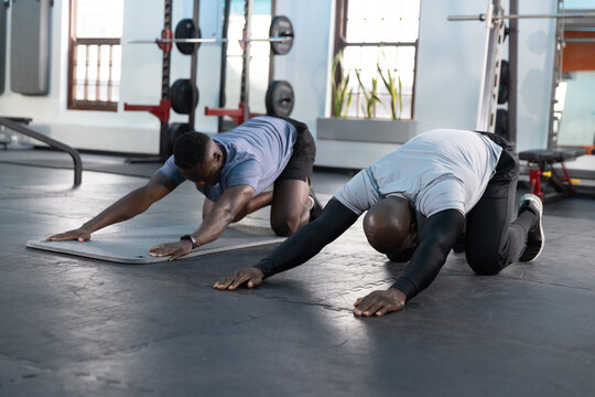 Two African American males performing deep kneeling forward stretches on mats in gym with barbells