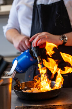 Sc&egrave;ne dynamique d'un chef en tablier noir utilisant un chalumeau de cuisine bleu pour flamber des aliments, avec des flammes vives s'&eacute;levant d'une po&ecirc;le au c&oelig;ur d'une cuisine professionnelle.