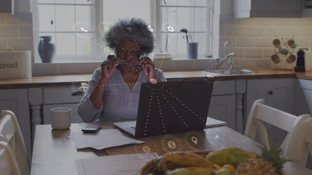 Senior woman fixing glasses at kitchen table, causing finance icons over laptop while calculating