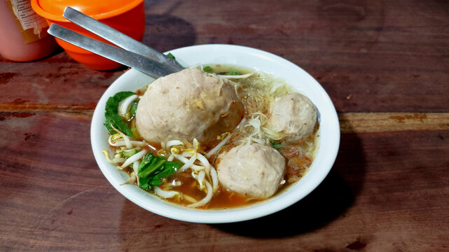 A bowl of traditional Indonesian bakso (meatball soup) served with noodles, bean sprouts, and greens in a savory broth. Captured on a rustic wooden table with a casual street food dining atmosphere.