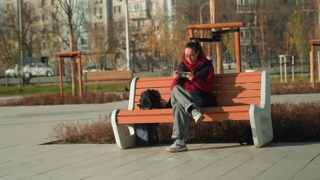 Woman on bench reading phone in park, student absorbed in message, red jacket, backpack at side, soft autumn light, empty plaza and concrete path, fallen leaves and contemplative mood