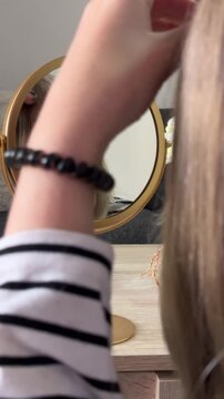 Focused teenage girl examining her face in a round vanity mirror, adjusting her blonde hair and bangs as part of her daily beauty and personal grooming routine at home