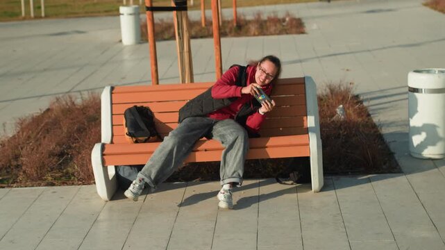 Woman seated relaxing with phone in hand. Overtired woman sitting quietly on bench in city environment. Fatigued female individual pauses on park bench under sun while engaging with her device