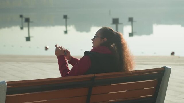 Caucasian woman on bench by waterfront scrolling phone in reflective mood, long hair haloed by backlight, distant boats and calm surface, serene golden light evokes solitude and quiet observation