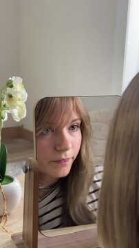 Unhappy teenage girl analyzing her face in a mirror, struggling with skin problems, low self-esteem, and body dysmorphia while looking at her reflection at home