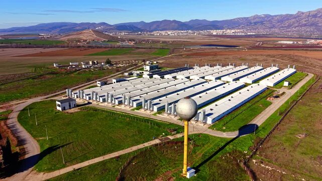 White agricultural buildings with metal silos and a spherical water tower in a rural landscape. Aerial view of large industrial poultry farm complex.