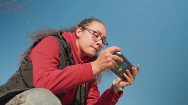 Young woman gaming on smartphone under blue sky, lowangle perspective highlights intense focus, fingers tapping touchscreen, red hoodie and black vest, sunny outdoor gameplay session on bench
