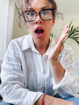 Woman wearing glasses and a white shirt reacting with a shocked, surprised, or amazed expression, holding her hand up while looking directly at the camera