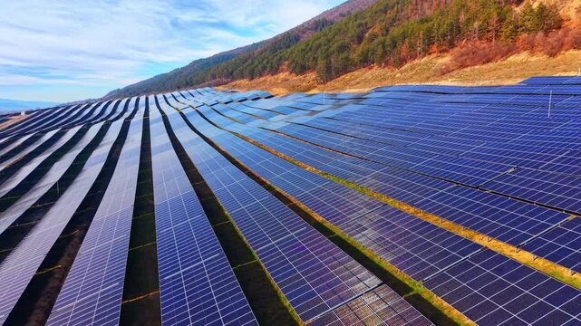 Aerial perspective long, uniform rows of photovoltaic solar panels installed on a grassy hillside slope. Geometric rows of solar panels.