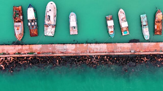 Concrete pier with tetrapods and fishing boats docked in clear water. Aerial view of fishing boats at pier.