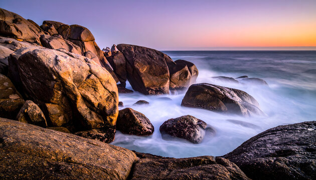 Coastal landscape at dawn with granite boulders and ocean waves smoothed into a soft white fog under a pink sky generative AI