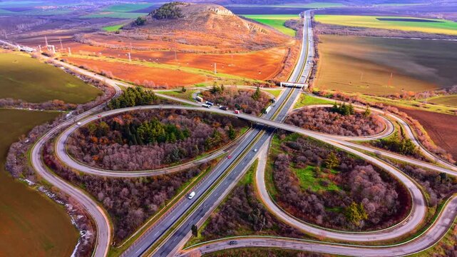 Road interchange. Vehicles are visible on the asphalt paths. Surrounded by autumn colored trees and farm fields. Symmetrical highway cloverleaf intersection.