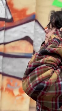 A young woman wears headphones while adjusting them with her hand. She is dressed in a colorful flannel shirt, highlighting her casual style in an urban setting.