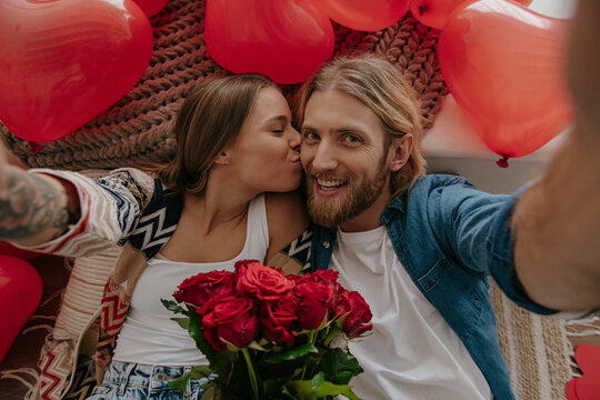 Top view of happy loving couple kissing and making selfie with red heart shape balloons around them