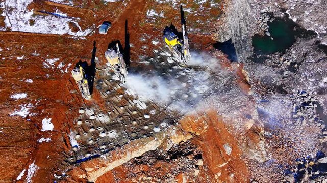 Top down aerial view of three heavy duty drilling machines creating holes in a rocky surface at a mining site with dust. Drilling rigs working on rock surface in open pit mine.