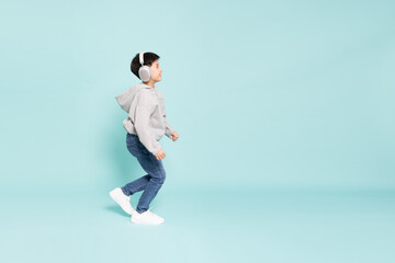 Fototapeta premium Happy young Asian boy wearing headphones and a gray hoodie walks joyfully against a teal background. Childhood music, active lifestyle, and modern technology