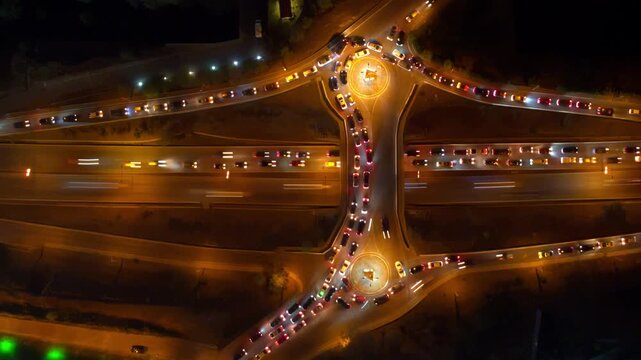 Latenight aerial ramp view in Tbilisi with flowing lanes and intermittent light trails, selective motion on curved ramps while adjacent lanes remain queued, amber streetlights sculpting road