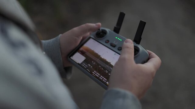 Close-up over-the-shoulder shot of a drone pilot's hands gripping the controller. The bright screen displays live aerial footage, with thumbnails and flight data visible, while thumbs hover over joyst