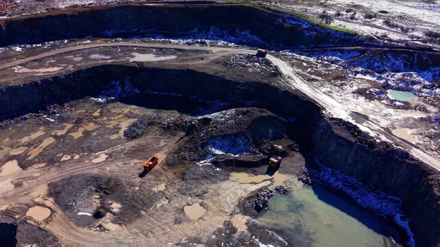 Multiple dump trucks and excavators moving earth and stone in a deep open pit mine with water accumulation at the bottom. Aerial view of mining operations in deep quarry.