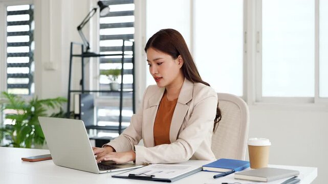 Focused asian woman using laptop, looking at screen, chatting, reading or writing email, sitting at desk, serious female working on research project online .