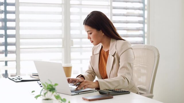 Focused asian woman using laptop, looking at screen, chatting, reading or writing email, sitting at desk, serious female working on research project online .