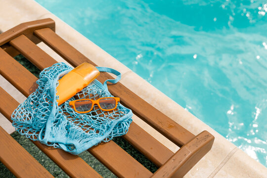 Summer essentials: sunscreen sunglasses and net bag on a wooden chair by a blue swimming pool