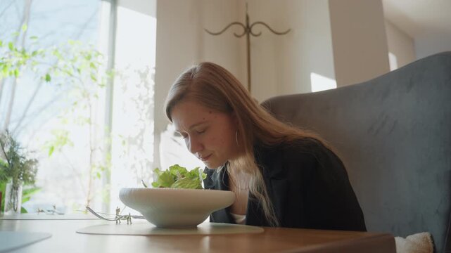 White woman executive at cafe table inspecting salad bowl with pensive expression, natural window light, cozy booth seating, slow deliberate sniff and pause before eating, subtle jewelry and blazer,