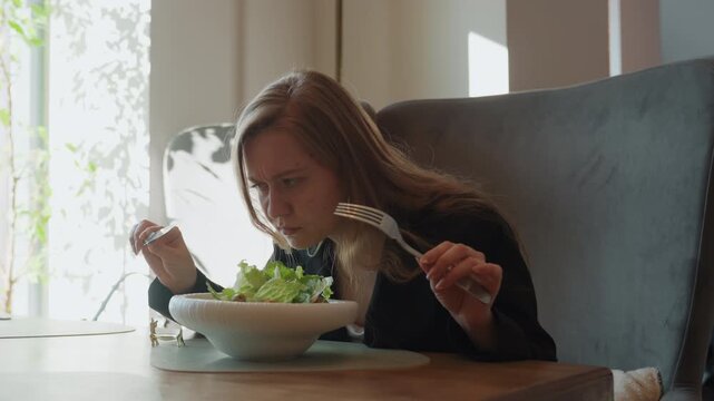 Caucasian woman reacting with disgust to salad, covers mouth and leans back in chair, troubled expression after sniffing bowl, suspicion of spoiled greens, interior cafe setting, natural light,