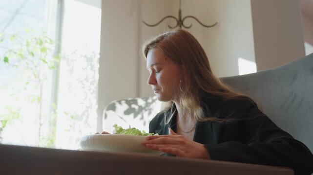 Young caucasian woman eating salad in sunlit cafe booth, wearing blazer and hoop earrings, focused on laptop and notes between bites, mindful slow meal with fresh greens on white plate, professional