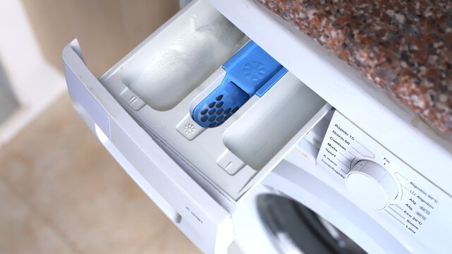 Washing machine detergent drawer with blue compartment for fabric softener and white sections for detergent, positioned on a granite countertop in a laundry room