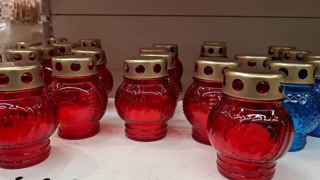 Row of red and blue memorial glass lanterns with golden metal caps in a shop display. Spiritual ritual attributes for grave decoration, cemetery commemoration, and Christian burial traditions