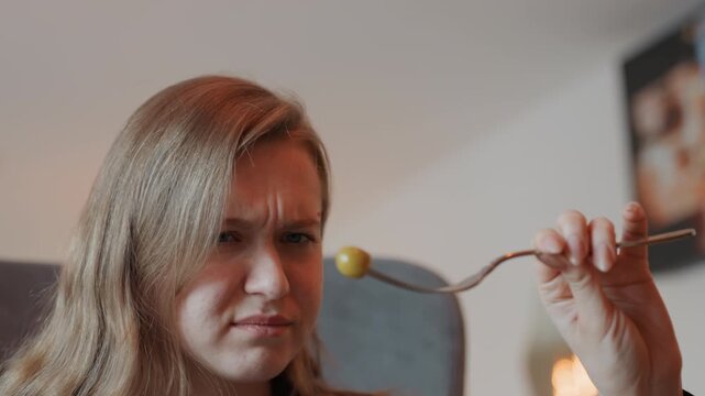 Young white woman tasting olive grimace inspecting forkful puzzled frown sour flavor shock covering mouth gag reflex candid home interior experimental taster uneasy aftertaste authentic reaction