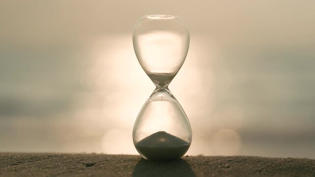 Glass hourglass on a sandy shore glowing in golden sunlight, representing time, waiting, deadlines, change, reflection, and the precious balance between past, present, and future.