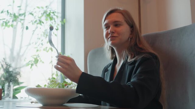 Caucasian woman tastes salad by window, remote worker sampling greens in cozy cafe, inspecting forkful, subtle smile, natural light, indoor plants, business casual blazer, warm cinematic tones